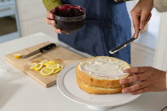 Two women wearing apron spreading frosting on layered cake, using offset spatula and jam on counter