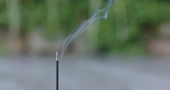 Smoldering incense stick is releasing curling smoke after ember ignition, scent drifting outdoors