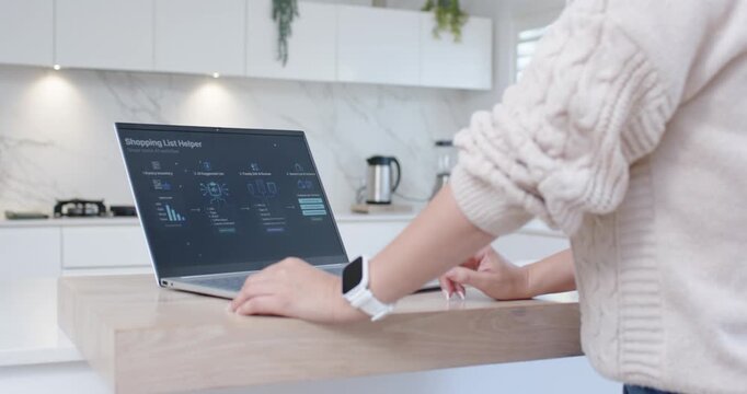 As laptop shows Shopping List Helper woman is tapping and updating list on wooden kitchen island