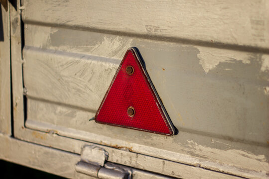 A red warning triangle is mounted on a gray metal surface. The sun shines brightly, illuminating the details of the triangle. The scene shows a simple outdoor environment with no distractions