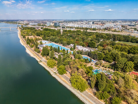 Aerial view of an outdoor lido and pools on Margaret Island beside the Danube River, surrounded by parkland and the Budapest cityscape, Hungary, in late summer.