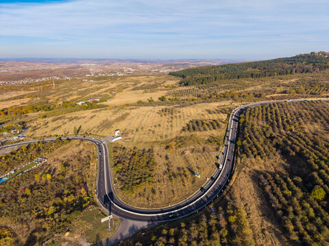 Aerial view of the winding Feleacu 311A road cutting through the hilly landscape with agricultural fields and scattered trees in Cluj-Napoca, Judetul Cluj, Romania.
