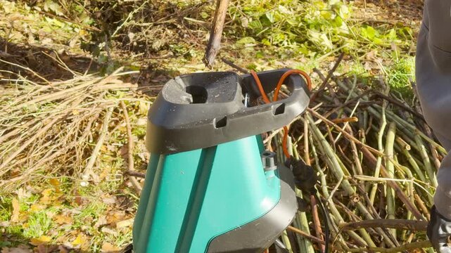 A gloved hand feeds a thick branch into an electric garden shredder in a grassy yard. Piles of cut branches surround the machine. The chipper makes a loud grinding sound.