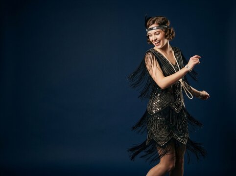 Laughing woman in 1920s flapper dress dancing against dark blue studio backdrop. Fringed sequin costume with pearl necklace and feather headband. Halloween party outfit.