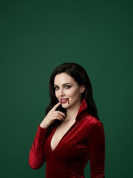 Vampire woman with blood on lip touching mouth in red velvet dress. Dark hair seductive look against green background. Halloween costume and vampire makeup concept.