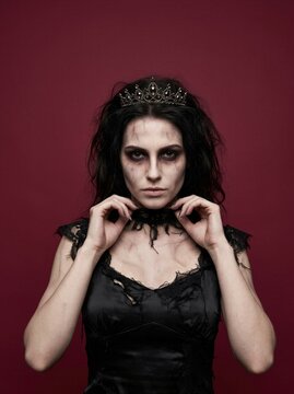 Zombie bride with tiara and torn black lace dress staring at camera on dark red background. Undead woman with scary makeup touching choker. Halloween horror costume.