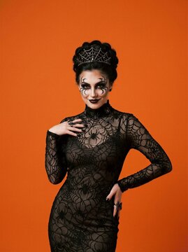 Woman in black spider web lace Halloween costume with tiara and spider face paint on orange background. Beauty portrait of dark queen makeup. Spooky costume party.