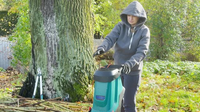 A person pushes sticks into a shredder beside a large tree trunk. The motor hums loudly, producing a harsh grinding and crunching sound.