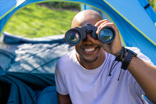 African man sitting at blue tent using black binoculars and smiling, wearing pink Tshirt, bracelets