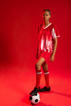 African-American woman standing in red studio with foot on soccer ball wearing red kit