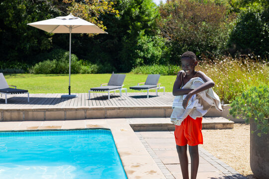 Adolescent African American male wearing red trunks standing at pool deck holding towel, copy space