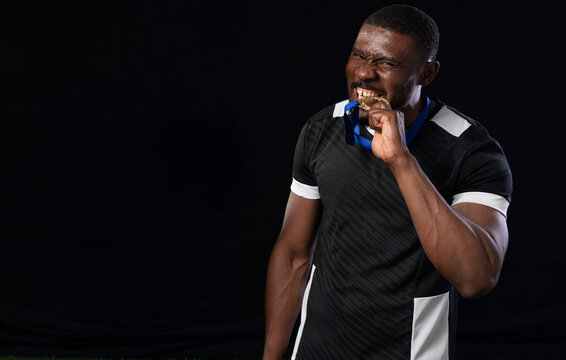 African man wearing black-and-white jersey biting gold medal on blue ribbon in studio, copy space