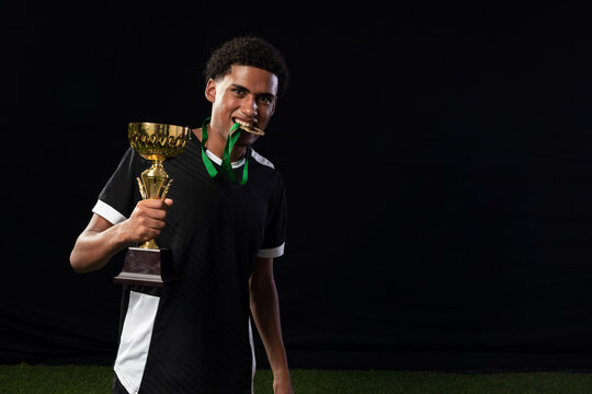 African American man in jersey holding gold trophy biting green-ribbon medal studio turf