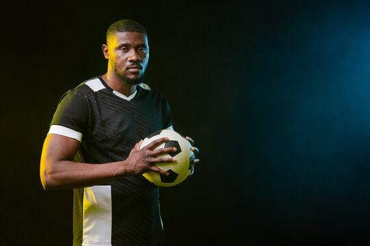 African American man holding soccer ball in studio, wearing black jersey, studio lights, copy space