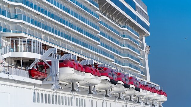 Cruise ship lifeboats, a row of red and white lifeboats on davits below many decks of balcony cabins on a large cruise ship, against a clear blue sky