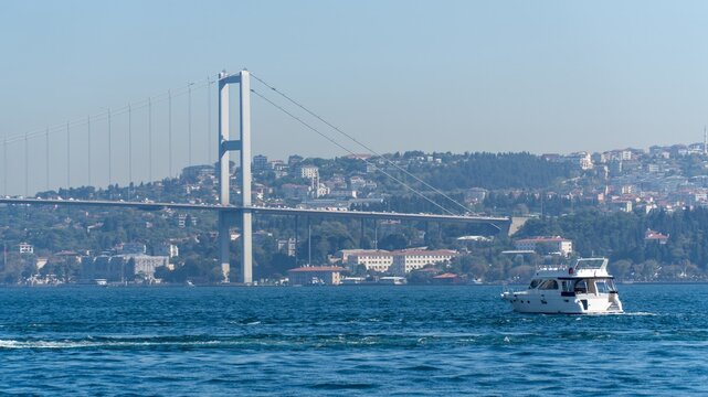 Bosphorus Bridge, the 15 July Martyrs Bridge, in Istanbul, Turkey, spanning the strait between Europe and Asia, with a white motor yacht cruising past the hillside