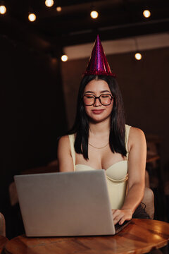 Beautiful woman wearing a birthday hat, using her laptop in a bar. Concept: working on one's birthday.