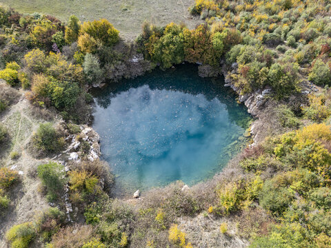 Aerial view of the Hordhanat e Virit karst spring with its deep blue water surrounded by vibrant autumn foliage and rocky terrain in Bajze, Shkoder County, Albania.