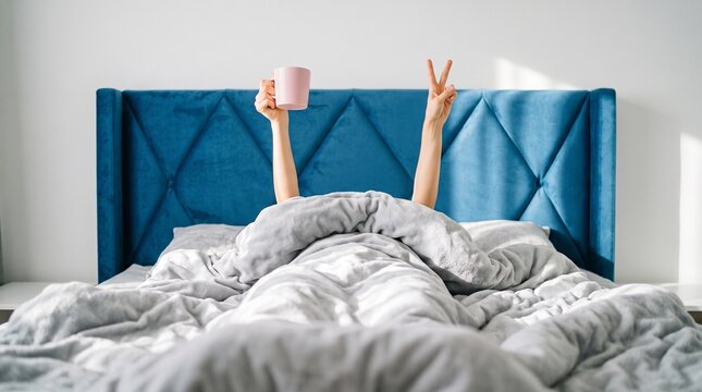 Person lying under a duvet in a cozy bed raises hands showing a peace sign and holding a coffee mug against a blue headboard.
