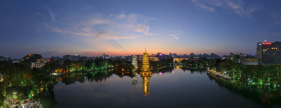 Aerial view of the illuminated Sun and Moon Pagodas reflecting in the calm waters of Shan Lake at dusk in Guilin, Guangxi, China.