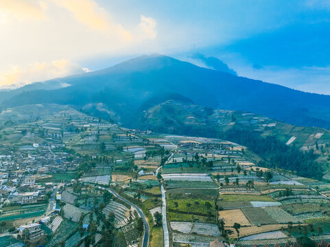 Aerial view of the terraced agricultural fields and village at the foot of Mount Sumbing with lush green slopes and golden hour light in Kecamatan Kaliangkrik, Jawa Tengah, Indonesia.