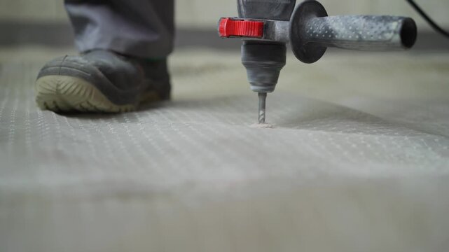 Close up of a builder drilling a hole in a textured flooring membrane during renovation or construction. The camera focuses on the drill bit penetrating the surface, then the finished hole