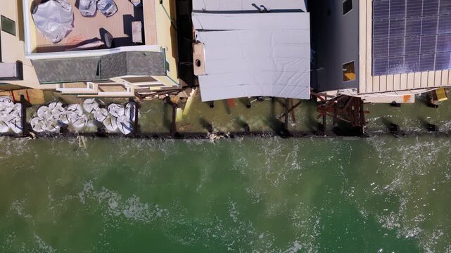 Long Beach, California, USA - October 15, 2026: Top down flight right to left over storm damaged, tarped homes protected by rows of sandbags along the broken bulkhead after Tropical Storm Rachel.
