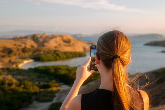 Komodo, East Nusa Tenggara, Indonesia - 26 July 2026: View of a woman taking a photo of the landscape at Komodo National Park with golden hour light illuminating the hills and water.