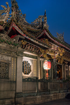Taipei City, Taiwan - 29 April 2015: View of the ornate roof and traditional architecture of Longshan Temple at dusk, with intricate stone carvings, gold details, and a red lantern.