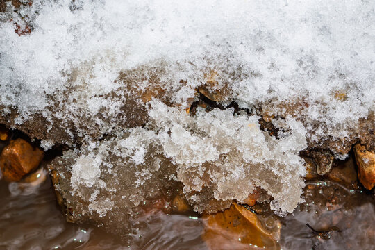 Crusty melting snow sitting atop frozen brown soil and wet pebbles, capturing the raw transition from winter to early spring near a flowing brook in March forest landscape