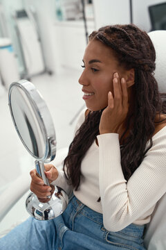 African woman beams with confidence while holding a round vanity mirror in a cosmetology clinic, appreciating a refreshed appearance and smooth skin after a beauty visit.