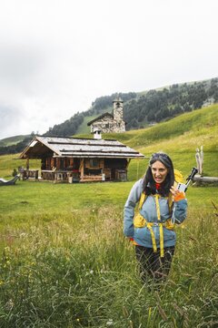 Disgusted woman hiker in waterproof clothing pulling funny face walking facing fears in tall high wet wildflower meadow background Malga Rossalm loop trail outdoor wilderness.Dolomites Italy