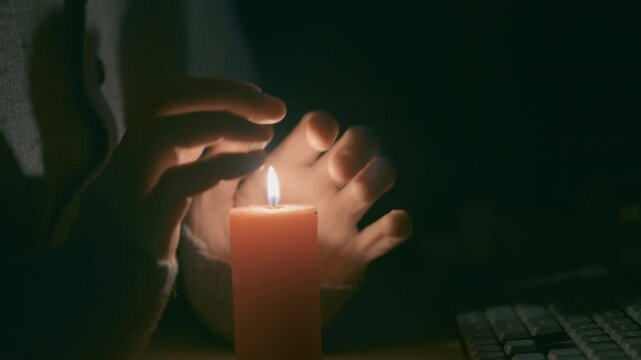 Man Warming Hands Over Burning Candle at Office Desk During Power Outage | Blackout Concept Man in Wool Sweater Near Computer Keyboard in Dark Room | Close-Up Shot | Energy Crisis and Winter Cold