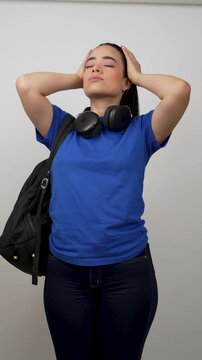Young latina woman with headphones and a backpack tying her long dark hair into a ponytail on a white background