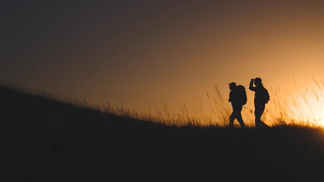 Business team silhouette hill. The two hikers walk together on a hillside, silhouetted by the sunset. The warm light guides the two hikers on their journey.