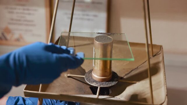 Close-up of metallic cylinder topped with dark powder being sampled with glass slide inside brass laboratory stand under warm light while gloved hand holds slide for inspection.