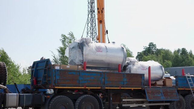 Crane lifting cylindrical stainless steel storage tanks wrapped in protective plastic onto flatbed truck at outdoor construction site, bright midday sky with trees in background