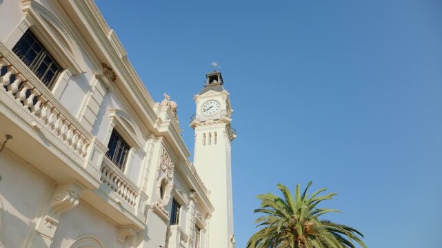 Sunlit historic clock tower and ornate stone facade with palm tree under clear blue sky during daytime pan movement, architectural detail and bright Mediterranean light