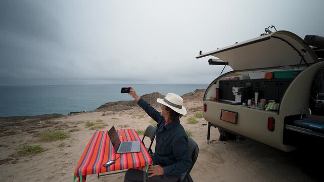 Mature man enjoys the outdoors by taking selfies and working on his laptop beside his teardrop camper during a peaceful off-grid retreat.