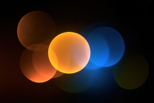 Glowing bokeh circles in orange, yellow and blue floating on black background, soft defocused lights for festive celebration and party design art