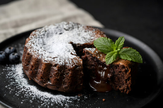 Chocolate fondant cake dusted with powdered sugar sits on a dark plate accompanied by fresh blueberries and a green mint leaf garnish.