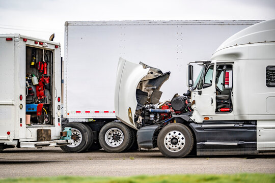 Broken white big rig semi truck with open hood standing on the industrial parking lot making repair with road help mobil assistant tech