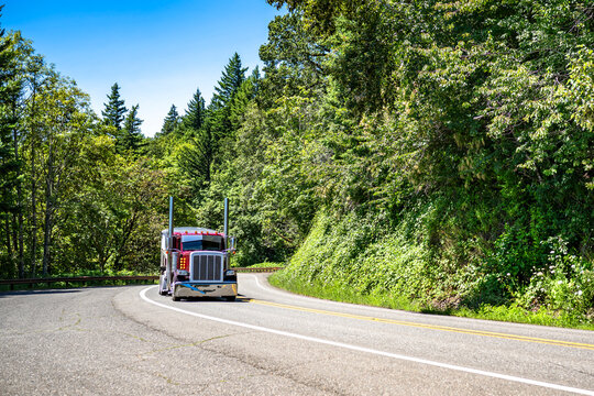 Burgundy classic big rig semi truck tractor with tall exhaust pipes transporting cargo in bulk semi trailer running on the winding mountain road in Columbia Gorge area