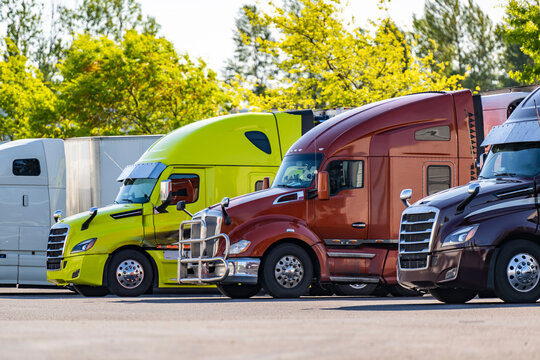Colorful big rig semi trucks with semi trailers standing in row on the truck stop parking lot with autumn trees on the back
