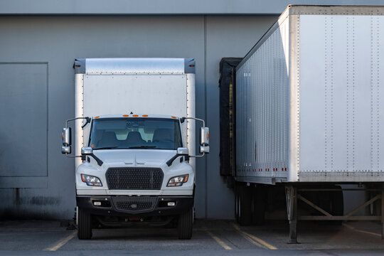 Front view of the day cab middle duty white rig semi truck with box trailer standing in warehouse dock gate beside with loading dry van semi trailer