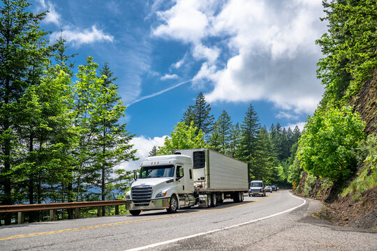 Low cab profile white big rig semi truck transporting cargo in refrigerator semi trailer running on the winding mountain road with rock wall and green trees