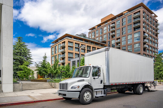 Middle duty local carrier day cab rig semi truck with box trailer and lowered lift system making delivery to customers standing on the urban city street with multilevel modern apartments