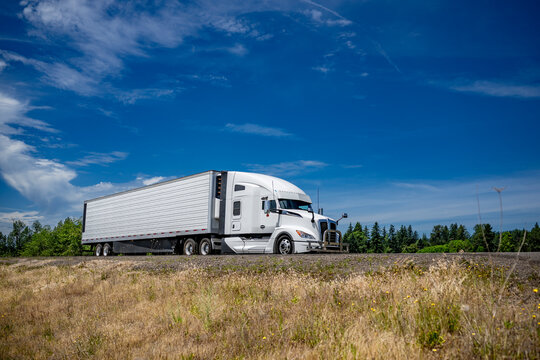 Powerful bonnet white big rig semi truck with grille guard transporting frozen cargo in refrigerated semi trailer running on the flat highway road with dry grass on the hill