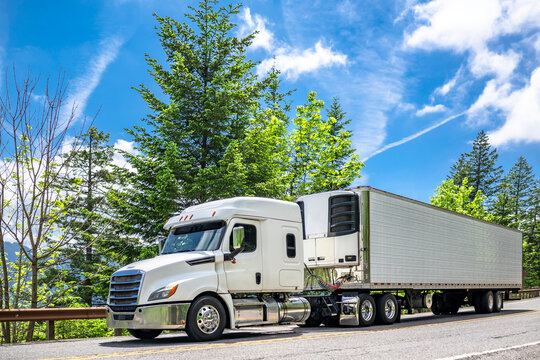 Powerful white big rig semi truck tractor with low profile extended cab transporting frozen cargo in refrigerated semi trailer driving on the mountain road with green trees on the side