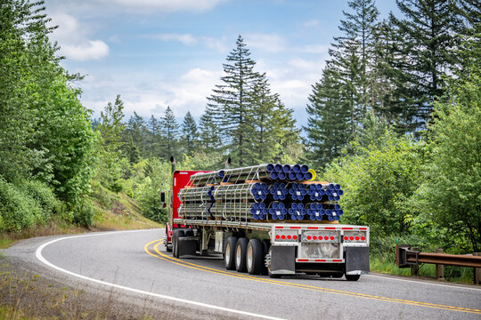 Red classic big rig semi truck transporting fastened pipes on flat bed semi trailer running on the winding road with green forest on the sides
